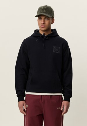 Jeune homme portant un sweat à capuche noir avec une poche avant, un pantalon bordeaux texturé et une casquette de baseball vert olive tournée vers l'avant.