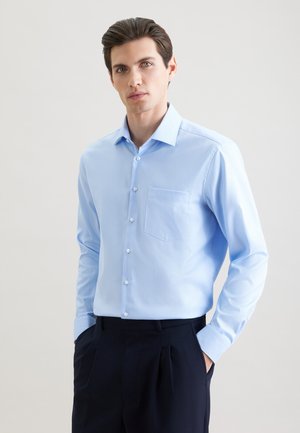 Jeune homme aux cheveux bruns portant une chemise bleu clair boutonnée et un pantalon foncé, les mains dans les poches, debout devant un fond uni.