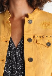 Detailní záběr na hruď a krk v žluté džínové bundě s kovovými knoflíky přes top s černo-bílým vzorem.