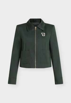 Femme debout portant une veste vert foncé avec un col gris, un pantalon large gris, des chaussures noires, tenant un sac à main noir.