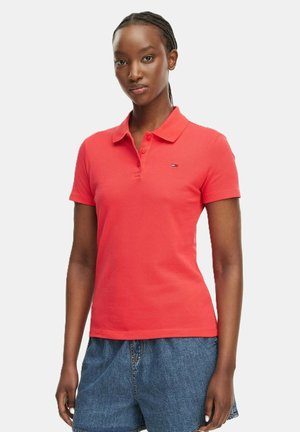 Mujer que lleva una camiseta polo coral de manga corta con un pequeño logo en el pecho y shorts vaqueros azules, de pie frente a un fondo blanco.