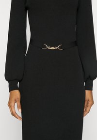 Femme portant une robe noire moulante côtelée avec de longues manches bouffantes et une fine ceinture noire avec une petite boucle dorée à la taille.