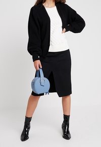 Musta neulottu neuletakki, valkoinen kaapelineulepaita, musta kynähame, jossa edessä halkio, kiiltävät mustat nilkkurit ja pyöreä sininen käsilaukku.