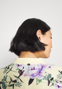 Floral blouse with a pastel yellow background, featuring purple and green floral patterns, a low collar, and a black button accent on the back.