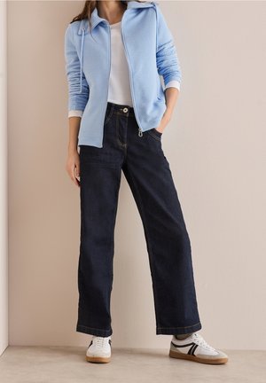 Person trägt eine hellblaue Jacke mit Reißverschluss, weißes Hemd, dunkle Jeans mit weitem Bein und weiße Sneakers mit schwarzen Streifen und steht an einer beigen Wand.