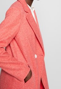 Blazer herringbone rosa con colletto standard, chiusura a un bottone e tasche laterali, realizzato in tessuto testurizzato.