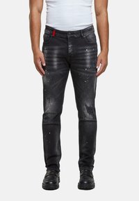Mustat farkut slim fit -mallissa, joissa on kulutettuja yksityiskohtia, maaliroiskeita ja punainen koristeellinen lenkki vyötäröllä.