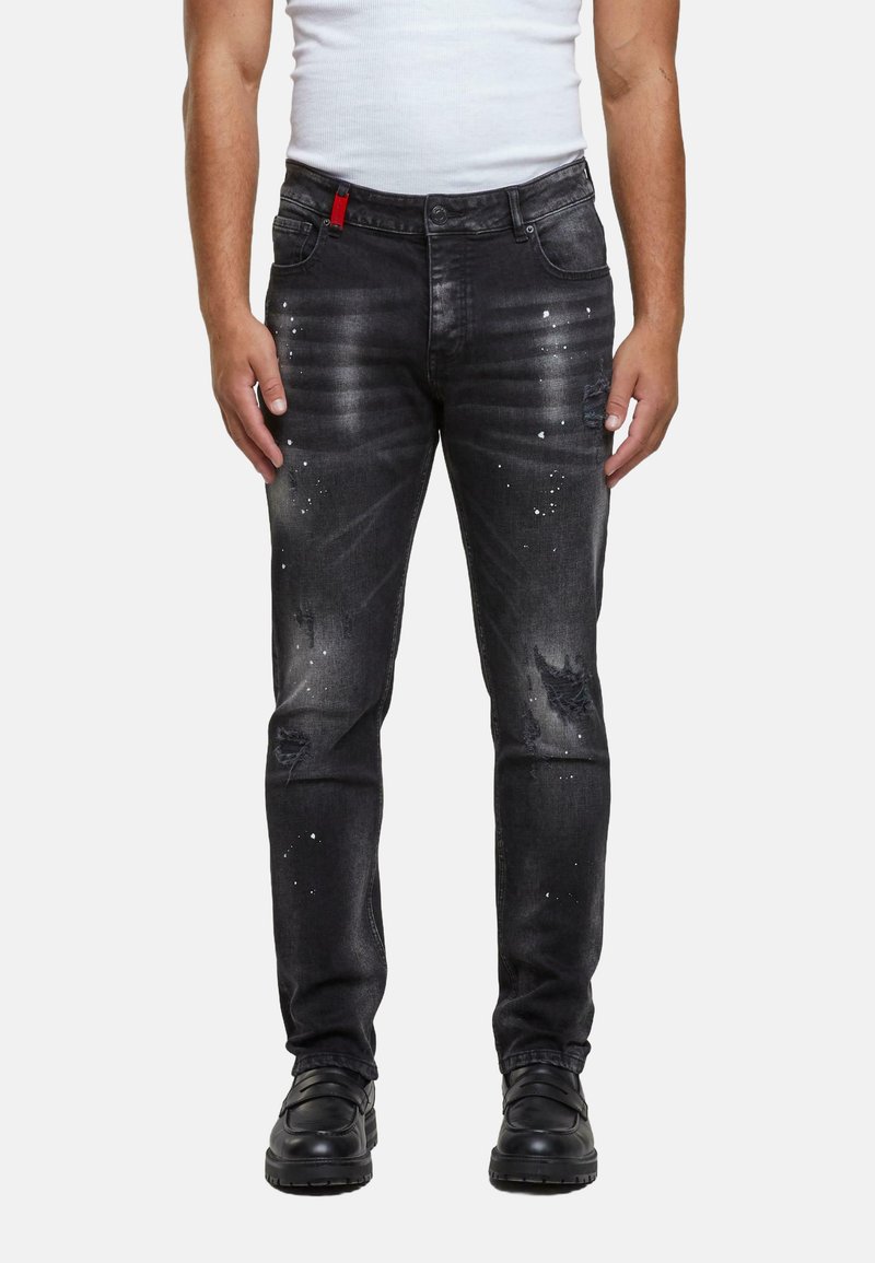 Jeans de denim negro con un corte slim, que presentan detalles desgastados, acentos de salpicaduras de pintura y un lazo decorativo rojo en la cinturilla.