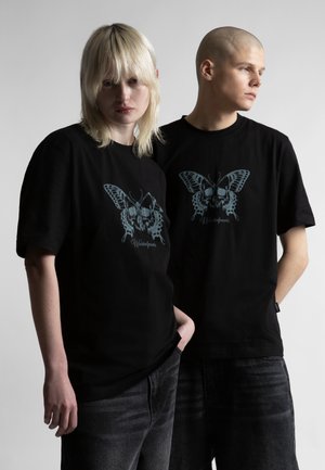 Dvaja mladí dospelí v čiernych tričkách s veľkým modrým motýľom a nápisom "Wasted Paris", stojaci pred jednoduchým pozadím.