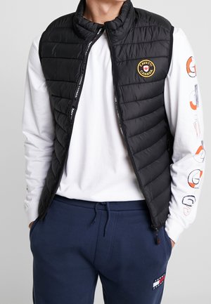 Gilet matelassé noir avec col montant, fermeture éclair et patch brodé. Porté par-dessus une chemise blanche à manches longues et un pantalon de survêtement bleu marine.