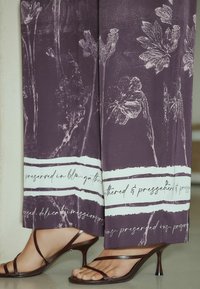 Lilla bukser med vide ben, hvide blomsterprint og kursiv tekst langs kanten, kombineret med mørkebrune sandaler med remme og lave kitten heels.