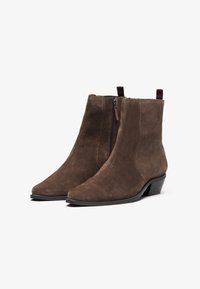 Botas de tobillo de ante marrón con punta afilada, cremallera lateral y un pequeño tacón cuadrado. Superficie texturada y lengüeta para facilitar el calzado.