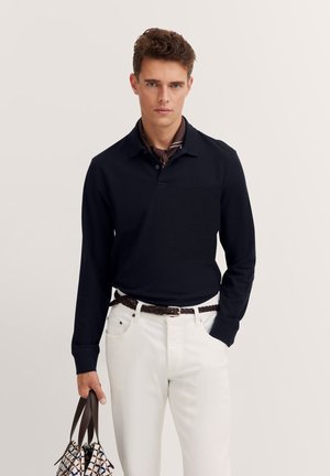 Navyblaues langärmliges Poloshirt in einfarbigem Design, mit Kragen und einer strukturierten Tasche. Getragen mit weißen Hosen und einer gemusterten Tasche.