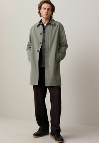 Pelerină de ploaie verde măsliniu cu un design ajustat, având guler clasic, închidere cu nasturi și buzunare laterale, asortată cu pantaloni închiși la culoare și pantofi.