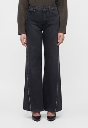 Person trägt schwarze Jeans mit hohem Bund und weitem Bein mit seitlichen Nieten, einen braunen Pullover und schwarze spitze Absätze.