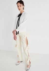 Blusa blanca con corbata negra, mangas largas; pantalones crema con aberturas laterales y lazos; bailarinas plateadas. Diseño minimalista, textura suave.