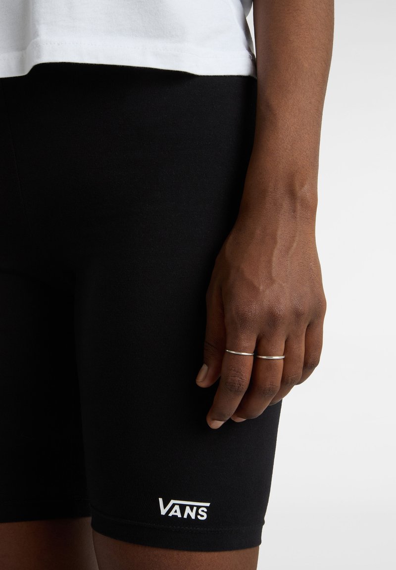 Pantalones cortos elásticos negros con un diseño ajustado, que presentan un logo blanco de "VANS" en la parte inferior de la pierna. Mano visible con anillos descansando sobre el muslo.