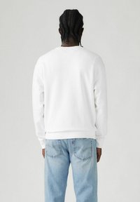 Sudadera blanca con puños y dobladillo acanalados, vista desde la parte trasera. Combinada con jeans de color azul claro que tienen bolsillos traseros. Textura suave.