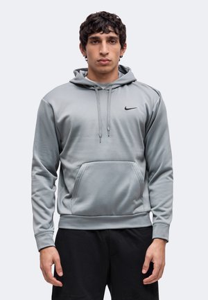 Jonge man staand, gekleed in een lichtgrijze Nike hoodie met voorzak en zwarte broek tegen een effen achtergrond.