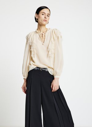 Blouse crème avec accents en dentelle volantée, encolure froncée et manches longues, associée à un pantalon noir à jambes larges et une ceinture fine.