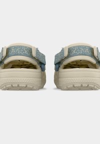 Sabots beiges avec des brides ajustables à motif bleu, semelles texturées et empeignes ventilées, vus de dos sur un fond blanc.