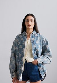 Denim jakna s starim vzorcem v modrih odtenkih, položena preko kreme barvane rebraste majice, v kombinaciji s temno modrimi kratkimi hlačami. Gladka tekstura.