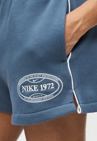 Κοντινό πλάνο σε μπλε σορτς Nike με άσπρο περίγραμμα, κεντημένο λογότυπο "NIKE 1972" και ένα χέρι στην δεξιά πλαϊνή τσέπη.