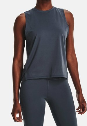 Vrouw draagt een donkergrijs sporttopje zonder mouwen en bijpassende leggings, staand met haar handen ontspannen langs haar zij.