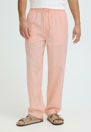 Mees, kes kannab heleroosa nööriga linaseid pükse ja beeže avatud varbaga sandaale, seisab valge tausta ees.