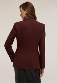 Donna con un blazer attillato di un intenso bordeaux scuro e una gonna nera, in piedi con le spalle rivolte verso la fotocamera, la testa girata di lato, e orecchini a cerchio dorati.