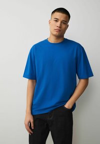 Jeune homme aux cheveux courts, portant un t-shirt oversize bleu vif uni et un pantalon noir, debout devant un fond gris clair.