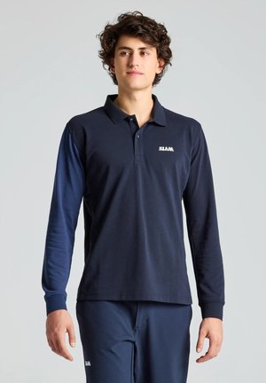 Un hombre luce un polo SELMA azul marino de manga larga y un pantalón a juego sobre un fondo liso y claro.