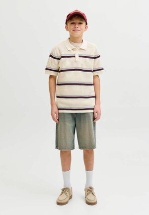 Chlapec v červenej čiapke, krémovo pruhovanej krátkorukávovej pletenej polokošeli, riflových šortkách, bielych ponožkách a béžových šnurovacích topánkach na jednoliatom pozadí.