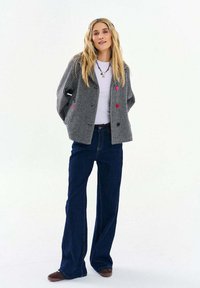 Žena s dlhými blond vlasmi, oblečená v čiernobielom saku s vzorom kacsacích zubov s farebnými gombíkmi, bielou košeľou, tmavomodrými džínsami s širokými nohavicami a hnedými topánkami.