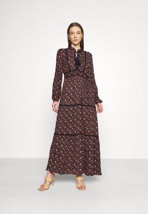 Robes maxi florales noires avec des manches longues, un nœud décoratif au niveau du col, des volants en couches, et une taille cintrée. Porté avec des sandales à talons beiges.