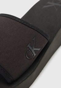 Sandale negre din material textil, cu o curea texturată având un logo negru proeminent, pe o talpă mată neagră.