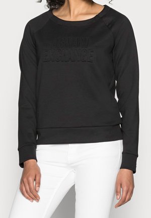 Femme portant un sweat-shirt noir à manches longues Armani Exchange avec logo embossé et pantalon blanc, représentée du cou aux hanches.