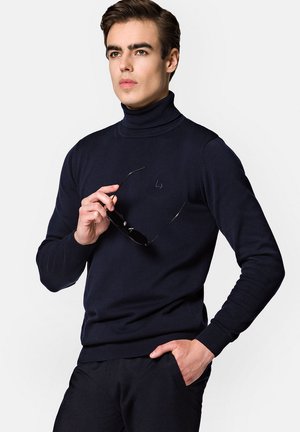 Granatowy sweter z golfem, wykonany z prążkowanej dzianiny, z małym haftowanym logo na piersi. Model trzyma okulary przeciwsłoneczne, prezentując dopasowany krój.