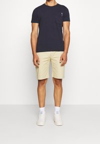 Námořnická modrá tričko s krátkým rukávem a přední kapsou, spárované s béžovými šortkami. Bílé tenisky s texturovanými podrážkami a bílé ponožky.