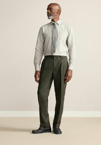 Hellgraues, strukturiertes Hemd mit Brusttasche; graue gestreifte Krawatte; dunkelgrüne, gefaltete Hose; schwarze Slipper. Einfaches, figurbetontes Design.