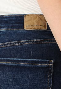 Sötét kék farmernadrág barna bőr címkével, amelyen az "AMERICAN EAGLE" felirat szerepel. Finom varrási részletekkel és sima felülettel rendelkezik.