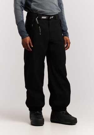 Persoon draagt zwarte sneeuwbroek met elastische enkels, zwarte winterlaarzen en een getextureerd grijs longsleeve, staand tegen een effen achtergrond.