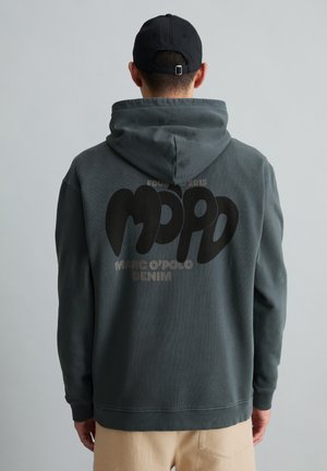 Mann trägt dunkelgrauen Kapuzenpullover mit dem Schriftzug „MPPD Marc O'Polo Denim“ auf dem Rücken und schwarze Kappe und steht vor einfarbigem Hintergrund.