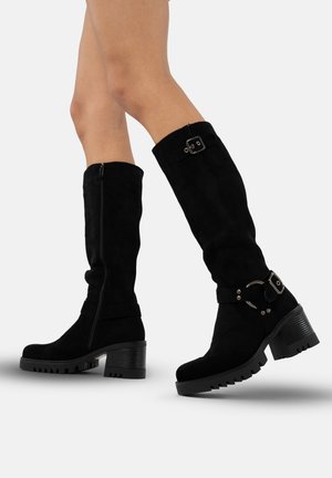 Bottes noires en suède jusqu'au genou avec un talon épais, présentant une boucle décorative et des détails de fermeture éclair, une semelle texturée et une finition lisse.
