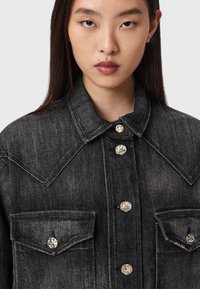 Jaqueta de ganga preta com colarinho clássico, botões prateados, dois bolsos frontais e detalhes de costura visíveis. Textura suave.