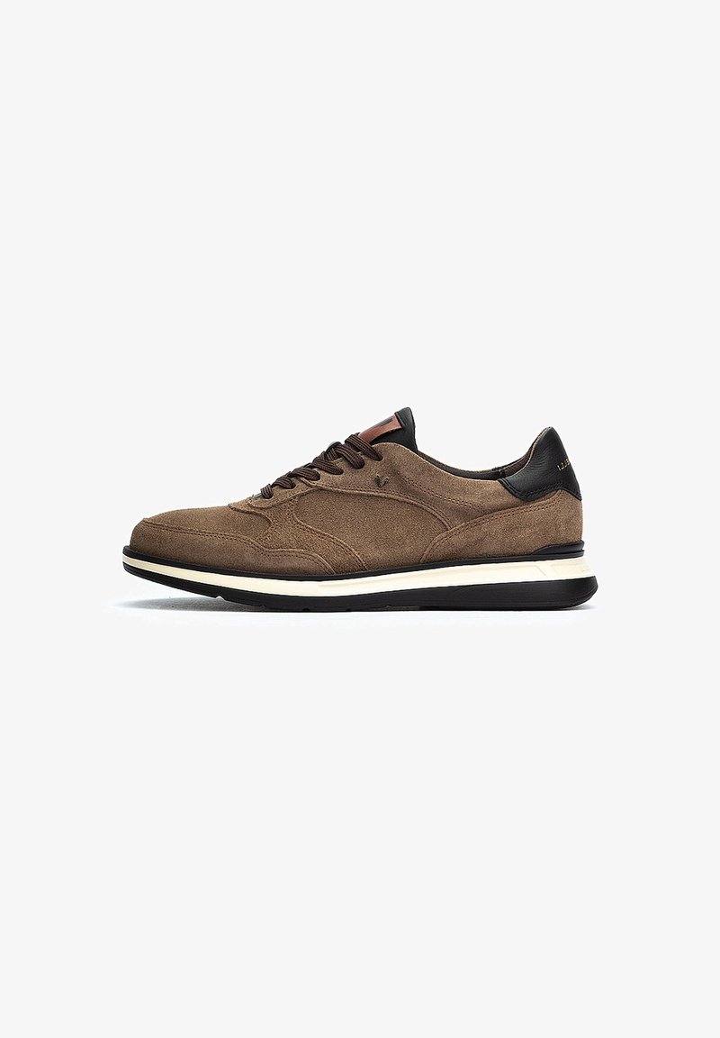 Zapatilla de ante marrón con un acento de cuero negro en el talón, que presenta cordones oscuros y una media suela blanca, textura suave y diseño atlético.