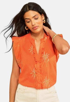 Femme aux longs cheveux foncés portant un chemisier sans manches orange à volants et imprimés de palmiers, associé à un pantalon clair et des bijoux en or.