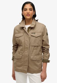 Beige utility jack met twee voorzakken, een hoge kraag en opgerolde mouwen met een groene accenten. Beschikt over een merkpatch op de arm.
