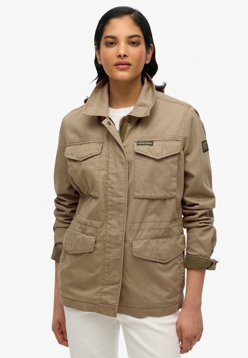 Beige utility jack met twee voorzakken, een hoge kraag en opgerolde mouwen met een groene accenten. Beschikt over een merkpatch op de arm.
