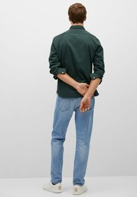 Green collared shirt with rolled sleeves, paired with light blue jeans and white sneakers. Man stands with hands behind back, viewed from the rear.